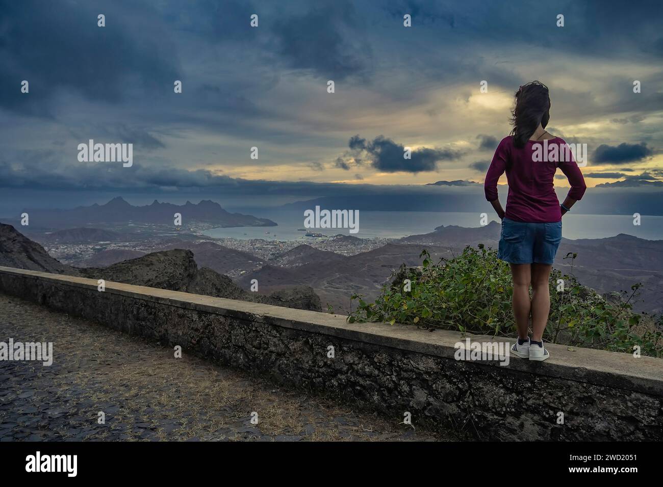 Una ragazza contemplativa si trova in un punto panoramico che si affaccia sulla città di Mindelo, São Vicente, Capo Verde, con uno skyline spettacolare sagomato contro una dinna Foto Stock