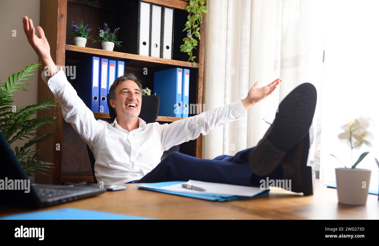 Uomo felice a braccia aperte seduto nel suo ufficio con i piedi sul tavolo di legno con documenti e scaffali con armadietti dietro Foto Stock