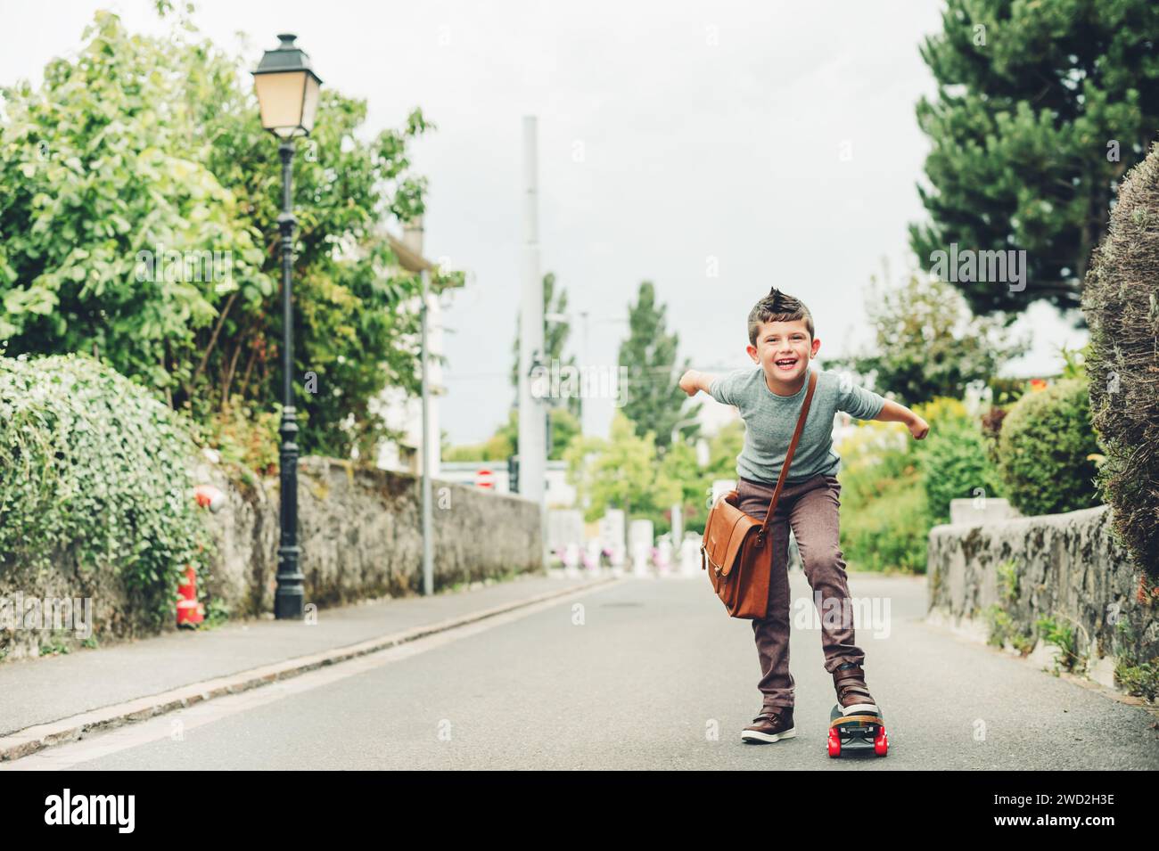 Ritratto all'aperto di un simpatico ragazzino che indossa una borsa in pelle marrone a spalla e che guida lo skateboard. Concetto di ritorno a scuola. Immagine tonificata dell'aspetto della pellicola Foto Stock