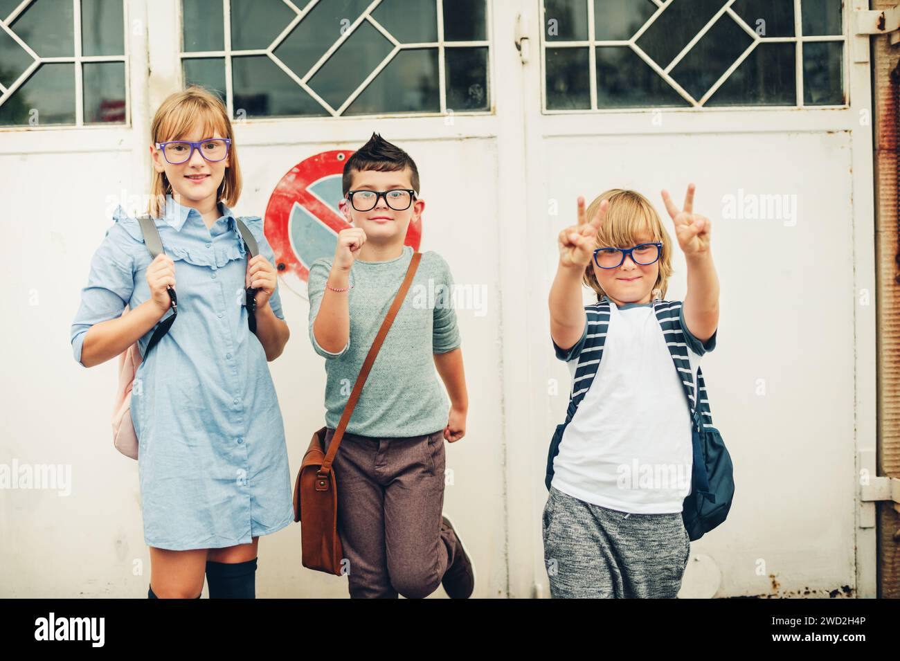 Gruppo di tre bambini divertenti che indossano zaini che tornano a scuola. Ragazze e ragazzi che indossano occhiali in posa all'aperto Foto Stock