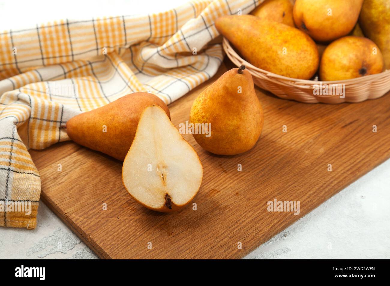 Tagliere in legno con interi e metà di succosa frutta di pera matura, cesto di vimini con pere e asciugamano da cucina giallo su fondo di legno bianco. Foto Stock