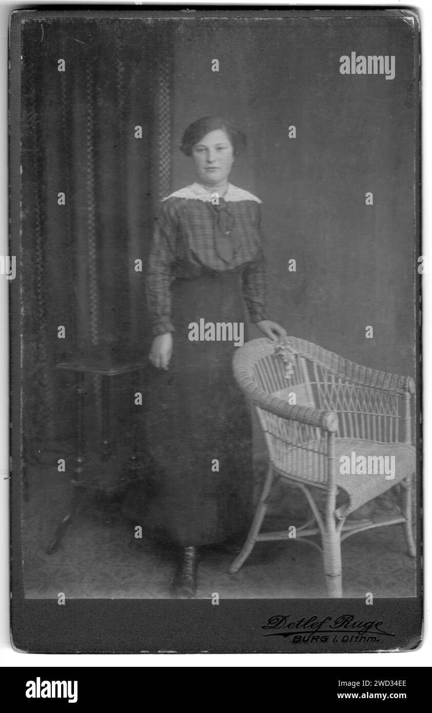 Ritratto in studio di una donna in una blusa a scacchi con colletto e cravatta in pizzo bianco, in piedi vicino a una sedia in vimini e un piccolo tavolo di legno, foto Foto Stock