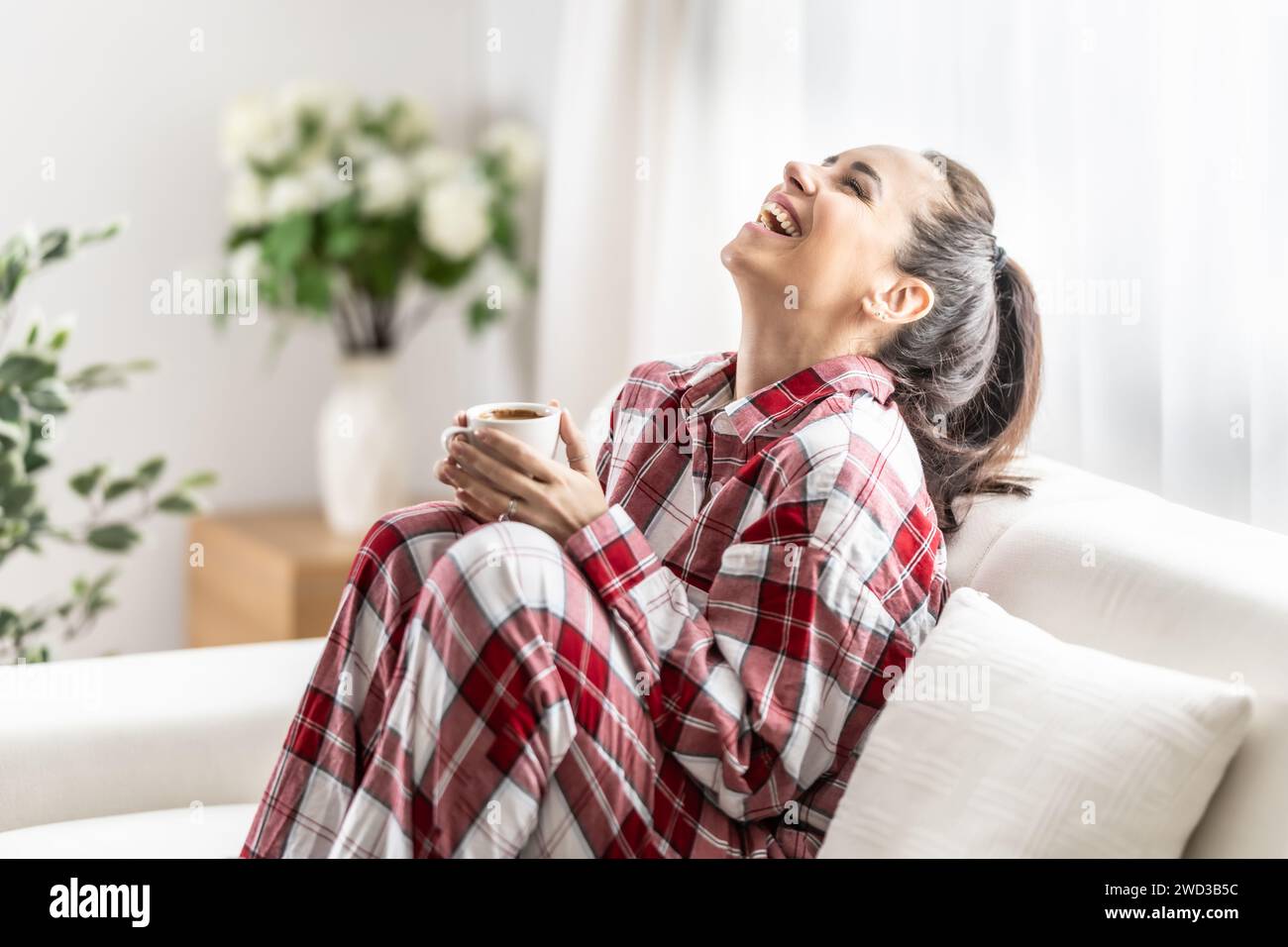 Giovane donna in pigiama rosso seduto sul divano e bevendo caffè al mattino in salotto, godendosi la giornata libera e ridendo. Foto Stock