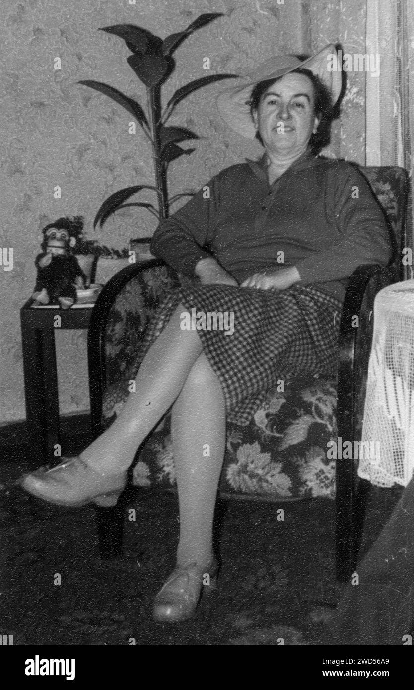 Una foto antica con mostra una donna di mezza età in una camicetta, un cappello e una gonna a pois, in posa, seduta su una sedia sullo sfondo di un albero di ficus Foto Stock
