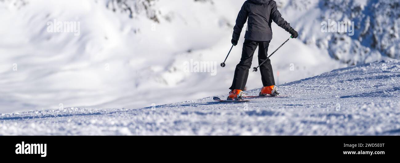 Uno sciatore, rivestito di una giacca scura e di scarponi di colore arancione brillante, intaglia un sentiero sulle scintillanti piste alpine, con le montagne innevate che offrono una maggiore maestosità Foto Stock