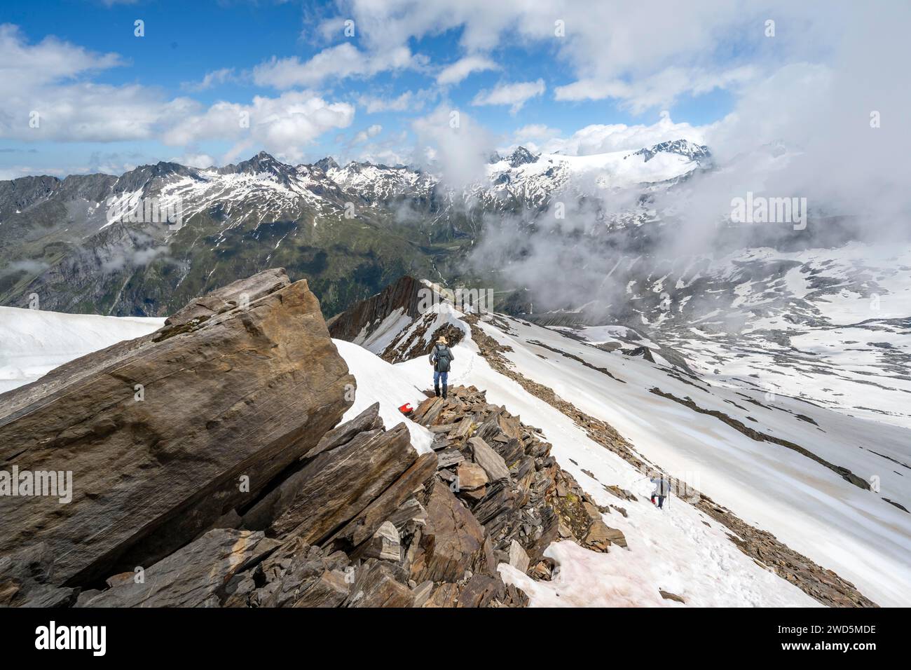 Alpinista su una cresta rocciosa con neve, discesa dalla cima del Corno di Schoenbichler, vista delle cime innevate e della valle Zemmgrund Foto Stock