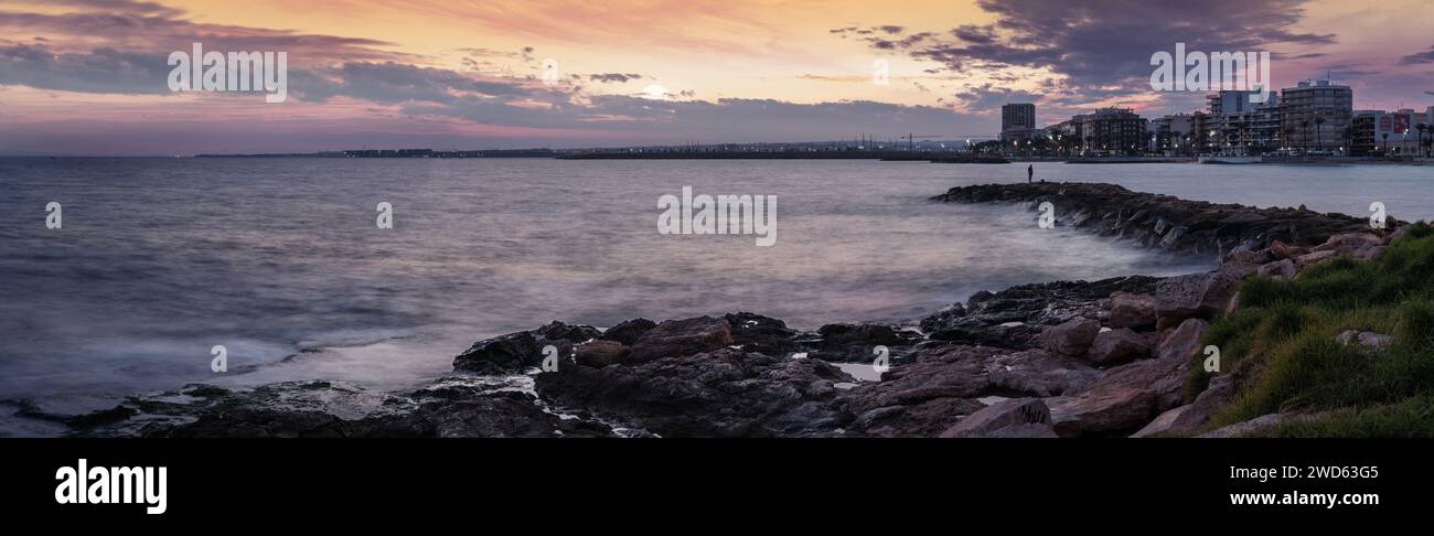 Torrevieja, Alicante, Spagna. Dietro un unico pescatore sul frangiflutti, le luci della città di Torrevieja lampeggiano mentre il sole tramonta sul Mediterraneo Foto Stock