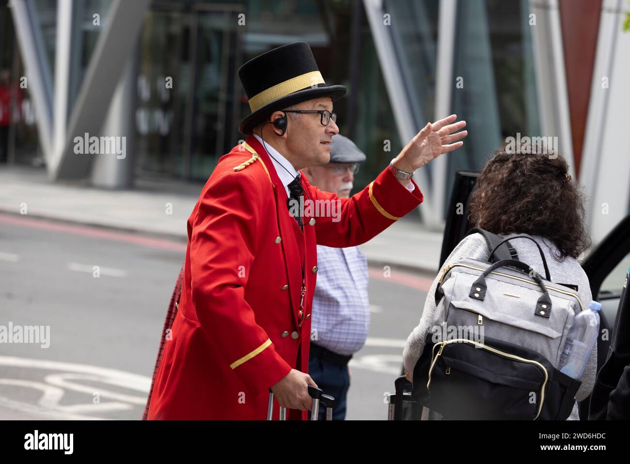 Un concierge vestito in elegante uniforme rossa tradizionale all'esterno del Rubens Hotel a Victoria, nel centro di Londra. Sta aiutando un passeggero a salire su un taxi nero Foto Stock