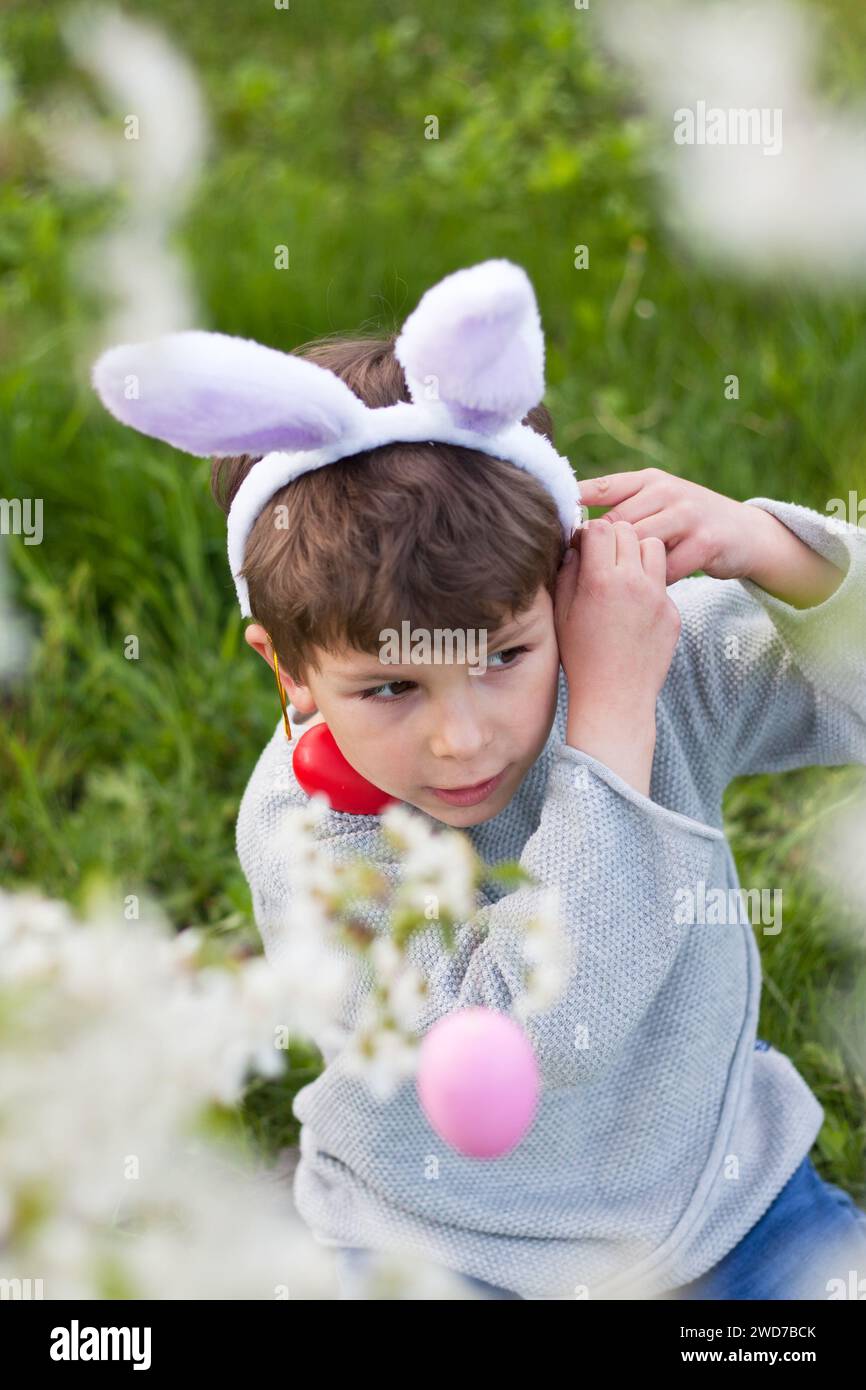 Caccia all'uovo di Pasqua. Bambino in età prescolare che indossa orecchie da coniglio per raccogliere uova colorate durante la caccia all'uovo di Pasqua nel giardino fiorito in primavera. Tradizione pasquale Foto Stock