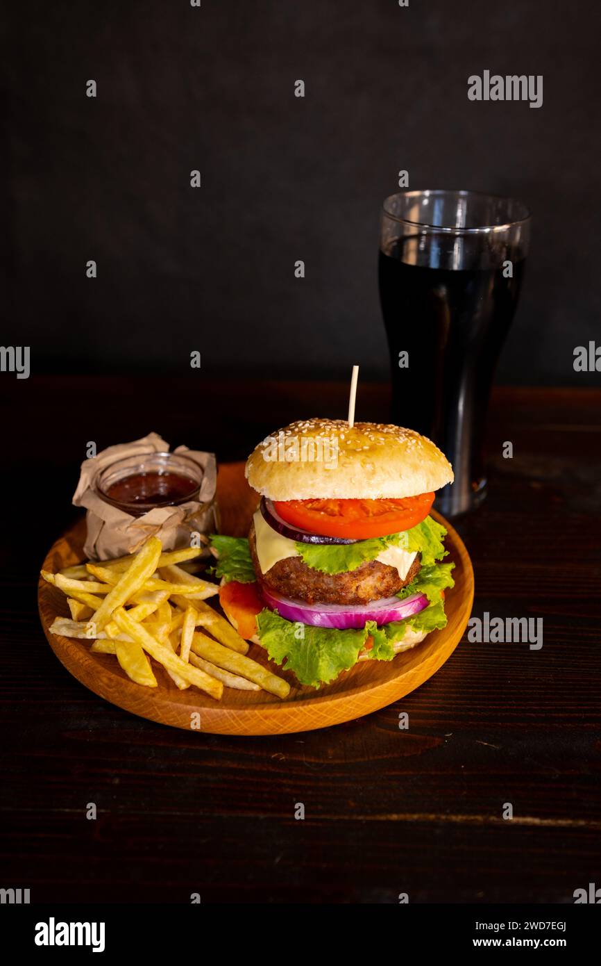 Hamburger delizioso con cola e patatine fritte su un tavolo di legno con fondo marrone scuro dietro. Concetto di fast food. Foto Stock