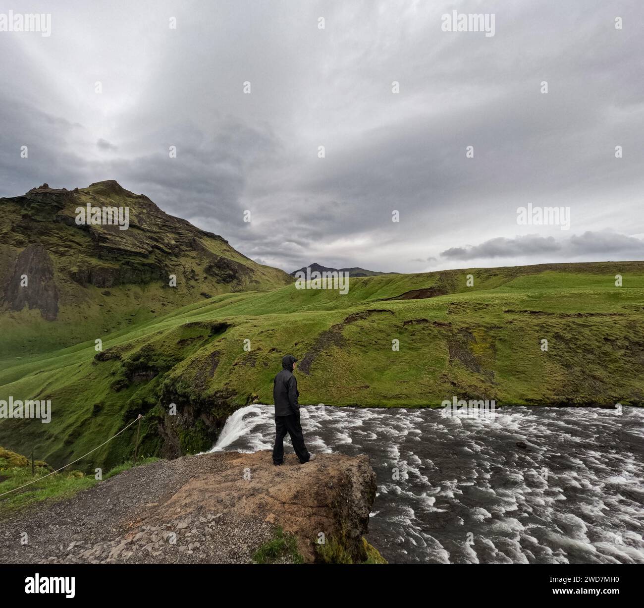 L'uomo cammina lungo il fiume con verdi colline erbose dietro il cielo nuvoloso Foto Stock