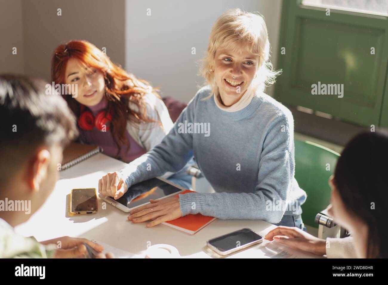 Un gruppo di studenti universitari allegri che collaborano utilizzando tablet e smartphone per la sessione di studio in una classe luminosa, che mostra interattti Foto Stock