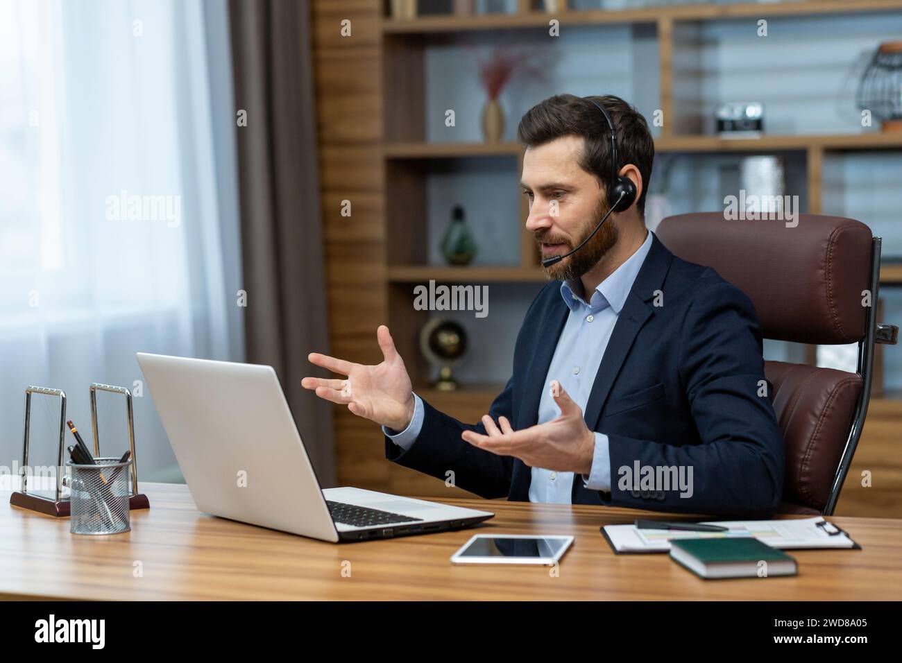 Uomo d'affari maturo con cuffie e auricolari impegnati in una chiamata in conferenza online mentre lavora in remoto da un ufficio domestico. Foto Stock