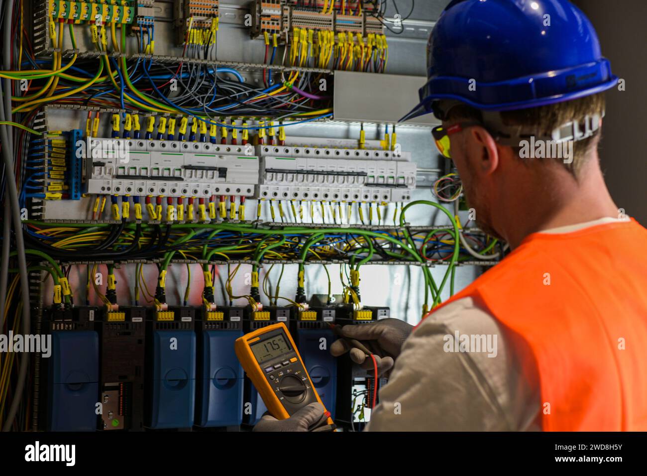 l'elettricista con elmetto blu, occhiali gialli e giubbotto arancione misura la corrente elettrica con multimetro digitale su sfondo sfocato nella scatola di distribuzione. Foto Stock