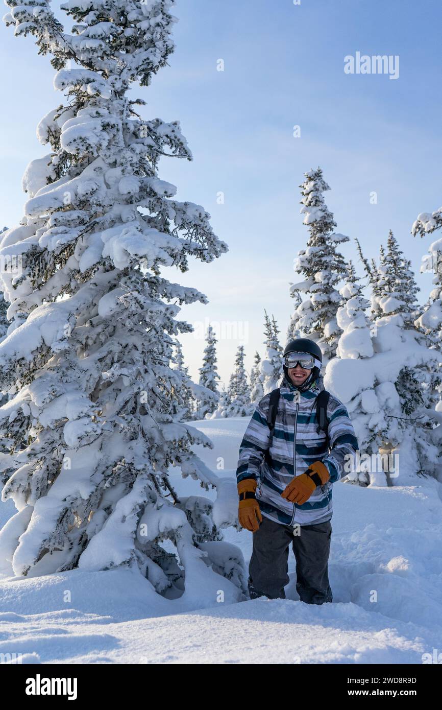 l'uomo sorridente si trova a fondo ginocchio nella neve vicino a un soffice albero di conifere innevato. Persona in equipaggiamento presso la stazione sciistica di Sheregesh, Siberia, Kemerovo Foto Stock