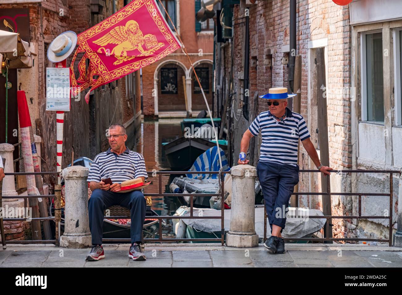 2018-07-03 Venezia, Italia. I gondolieri vecchi e giovani che indossano abiti tradizionali si trovano sul molo con la bandiera veneziana e i canali sullo sfondo Foto Stock