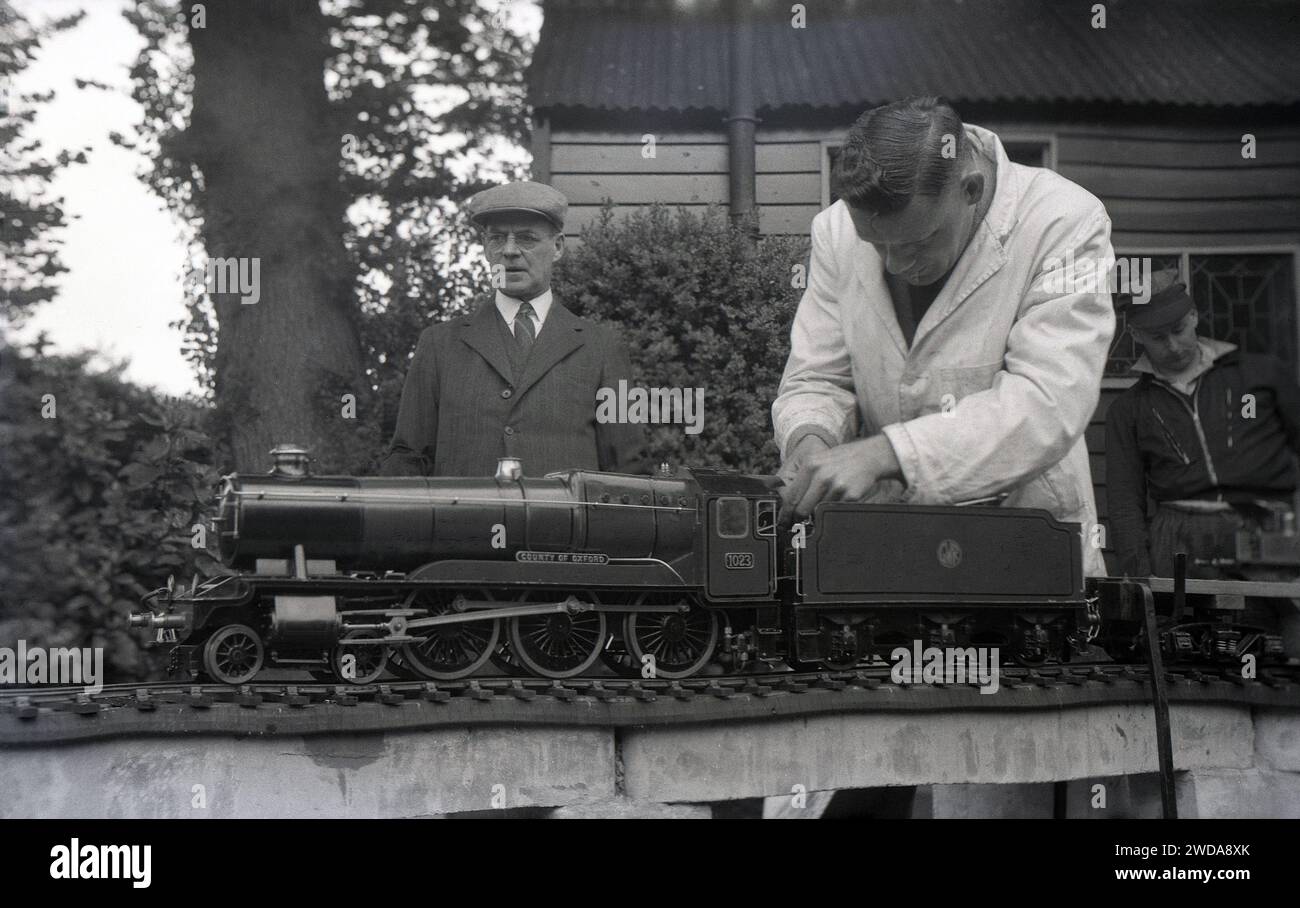 Anni '1950, storica, all'esterno nel giardino della Field End Railway Co, Inghilterra, Regno Unito, un modellino di ferrovia a vapore appassionato di un camice bianco che lavora su un treno a vapore in miniatura, 'County of Oxford' (1023) una locomotiva a vapore GWR che entrò in servizio nel 1947. La County Class o 1000 Class of Steam locomotive fu lo sviluppo finale della Saint Class a due cilindri originariamente introdotta nel 1901. Foto Stock