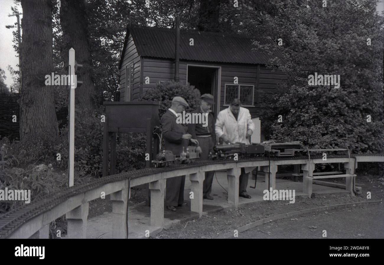 Anni '1950, storico, all'aperto in un giardino, appassionati di modellini di ferrovie , uno con un mantello bianco, accanto a una ferrovia a vapore in giardino in miniatura, presso la Field End Railway Co, Inghilterra, Regno Unito. Su un binario sopraelevato seduto su pali di cemento, il modello di treni a vapore, uno dei quali è un modello in scala della locomotiva a vapore GWR 'County of Oxford' (1023), il cui originale iniziò il servizio nel 1947. Foto Stock