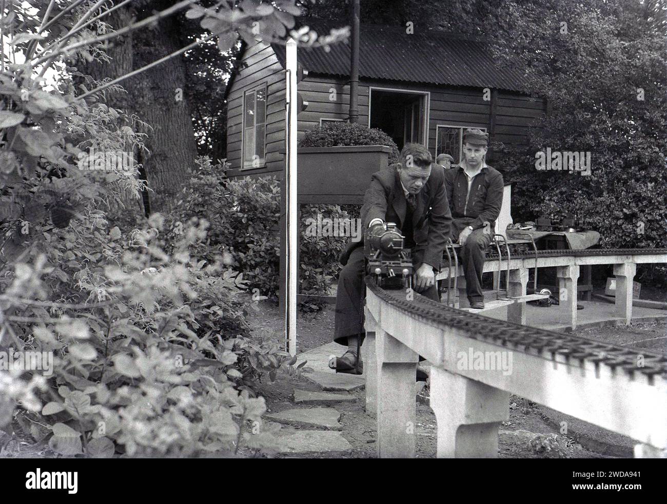 Anni '1950, storico, all'aperto in un giardino, appassionati di modellismo ferroviario a vapore in un giardino in miniatura, presso la Field End Railway Co, Inghilterra, Regno Unito. Su un binario sopraelevato seduto su pali di cemento, due uomini cavalcano sui minuscoli treni a vapore, uno dei quali è un modello in scala della locomotiva a vapore GWR 'County of Oxford' (1023 ), il cui primo è entrato in servizio nel 1947. Foto Stock