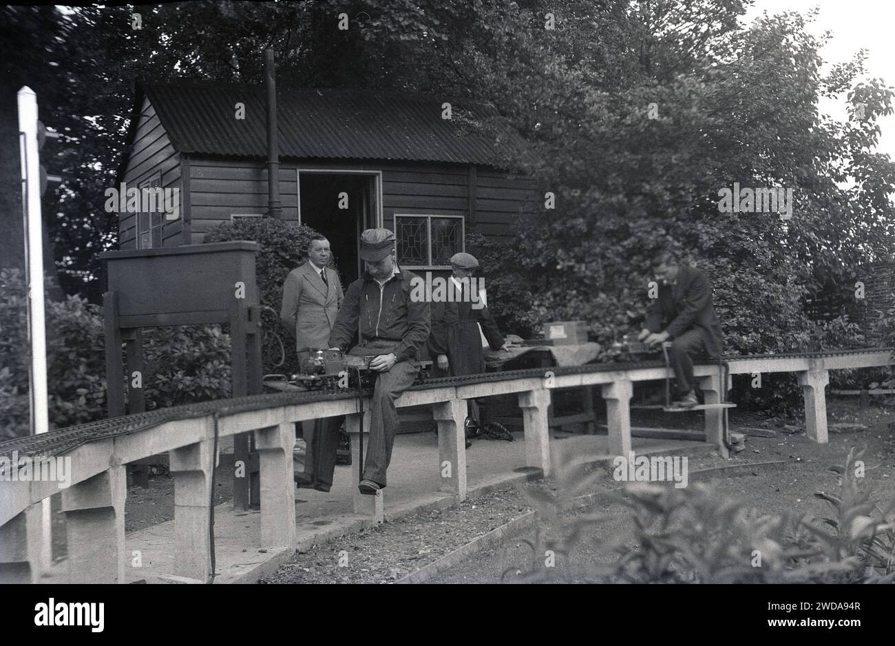 Anni '1950, storico, all'aperto in un giardino, appassionati di modellismo ferroviario a vapore in un giardino in miniatura, presso la Field End Railway Co, Inghilterra, Regno Unito. Su un binario sopraelevato seduto su pali di cemento, un uomo in giacca e berretto che cavalca un modellino di treno a vapore. Foto Stock