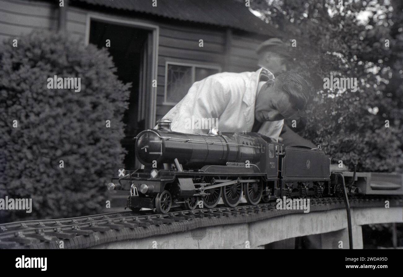 1950, storica, all'esterno nel giardino della Field End Railway Co, Inghilterra, Regno Unito, un modellino di ferrovia a vapore appassionato di un camice bianco che lavora su un treno a vapore in miniatura, 'County of Oxford', una locomotiva a vapore GWR entrata in servizio nel 1947. Foto Stock