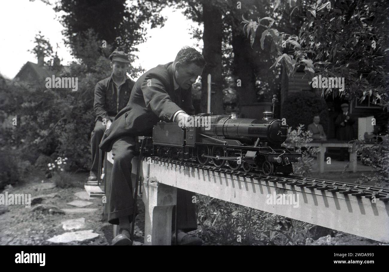 Anni '1950, storico, all'aperto in un giardino, appassionati di modellismo ferroviario a vapore in un giardino in miniatura, presso la Field End Railway Co, Inghilterra, Regno Unito. Su un binario sopraelevato seduto su pali di cemento, due uomini cavalcano sui minuscoli treni a vapore, uno dei quali è un modello in scala della locomotiva a vapore GWR 'County of Oxford' (1023 ), il cui primo è entrato in servizio nel 1947. Foto Stock