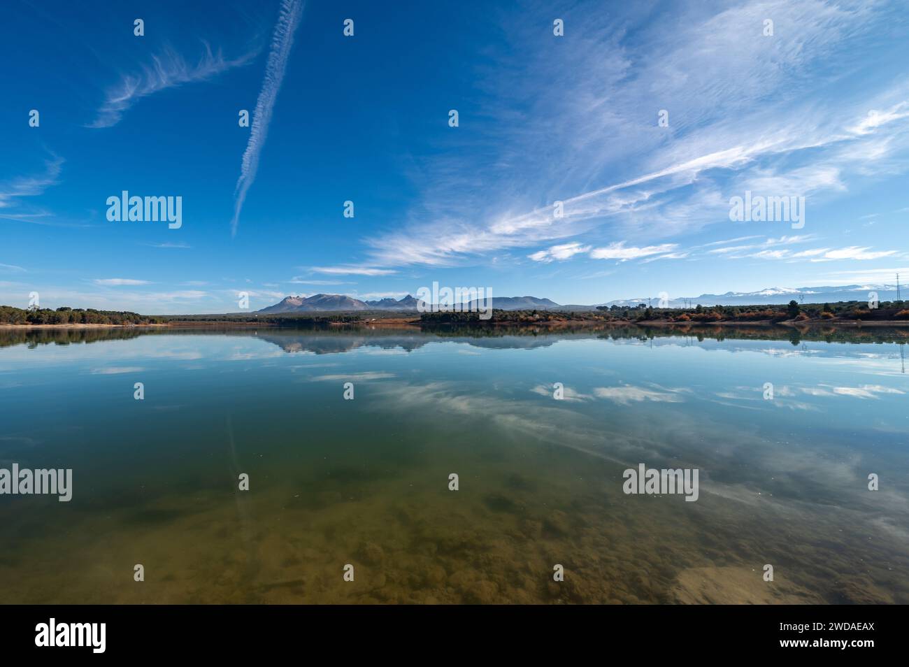 Riflesso di montagne e alberi in un bacino idrico di Granada (Spagna) in una soleggiata mattinata d'inverno Foto Stock
