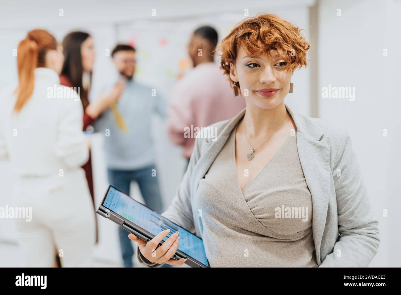 Donna d'affari incinta che tiene un laptop. Ritratto di donna d'affari. Persone che lavorano dietro. Foto Stock