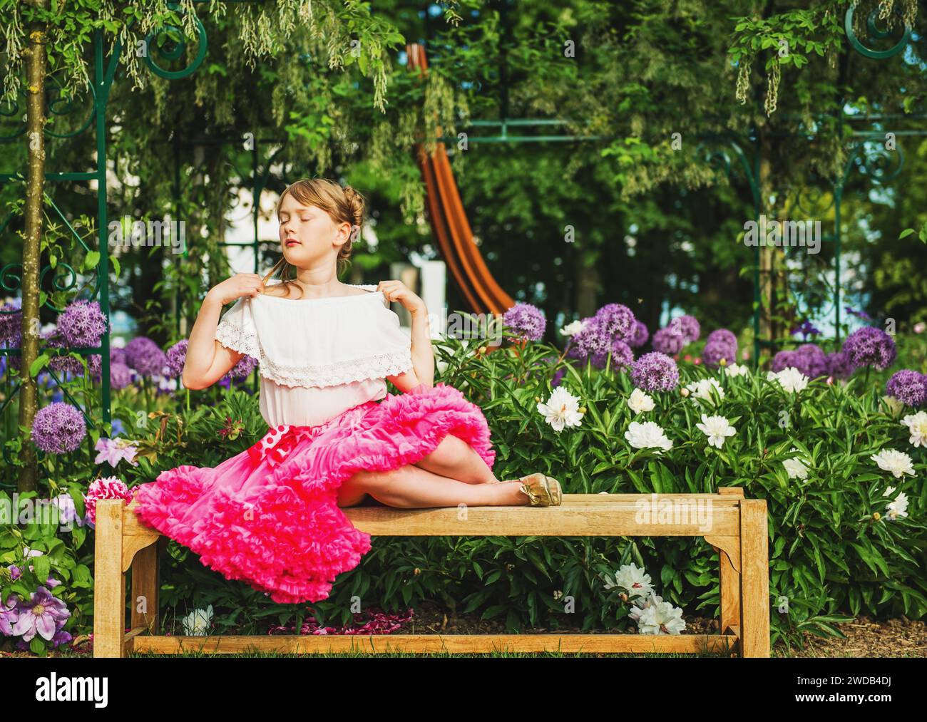 Simpatica bambina che riposa sulla panchina in un bellissimo giardino fiorito in una bella giornata estiva, indossa una camicia bianca e una gonna tutu rosa brillante Foto Stock