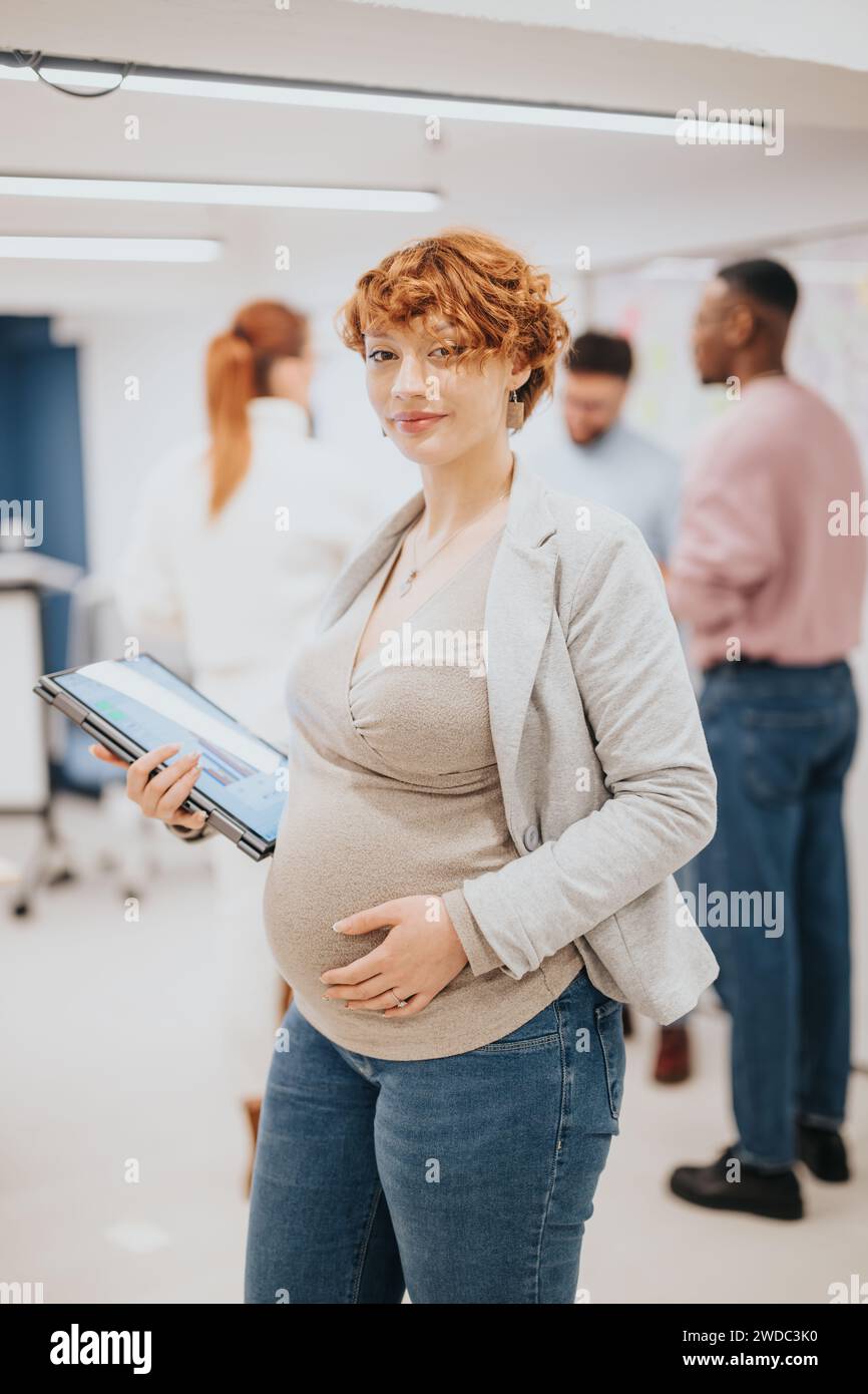 Splendida donna d'affari incinta rossa che si posa in ufficio mentre tiene il laptop. La sua squadra sta facendo il brainstorming alle spalle. Copia spazio. Foto Stock