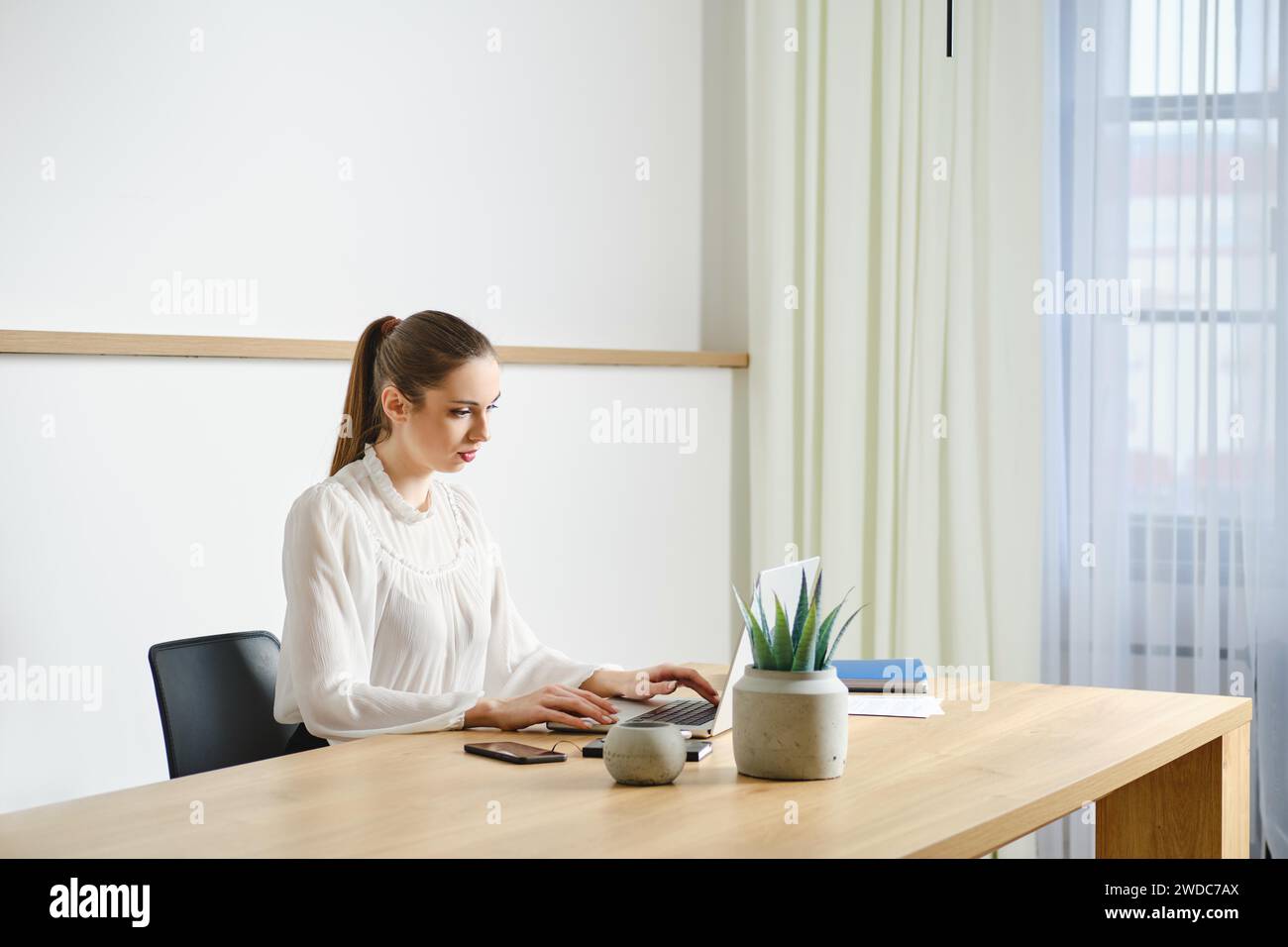 Una donna professionale concentrata lavora in una scrivania pulita e organizzata in un moderno ufficio ben illuminato Foto Stock