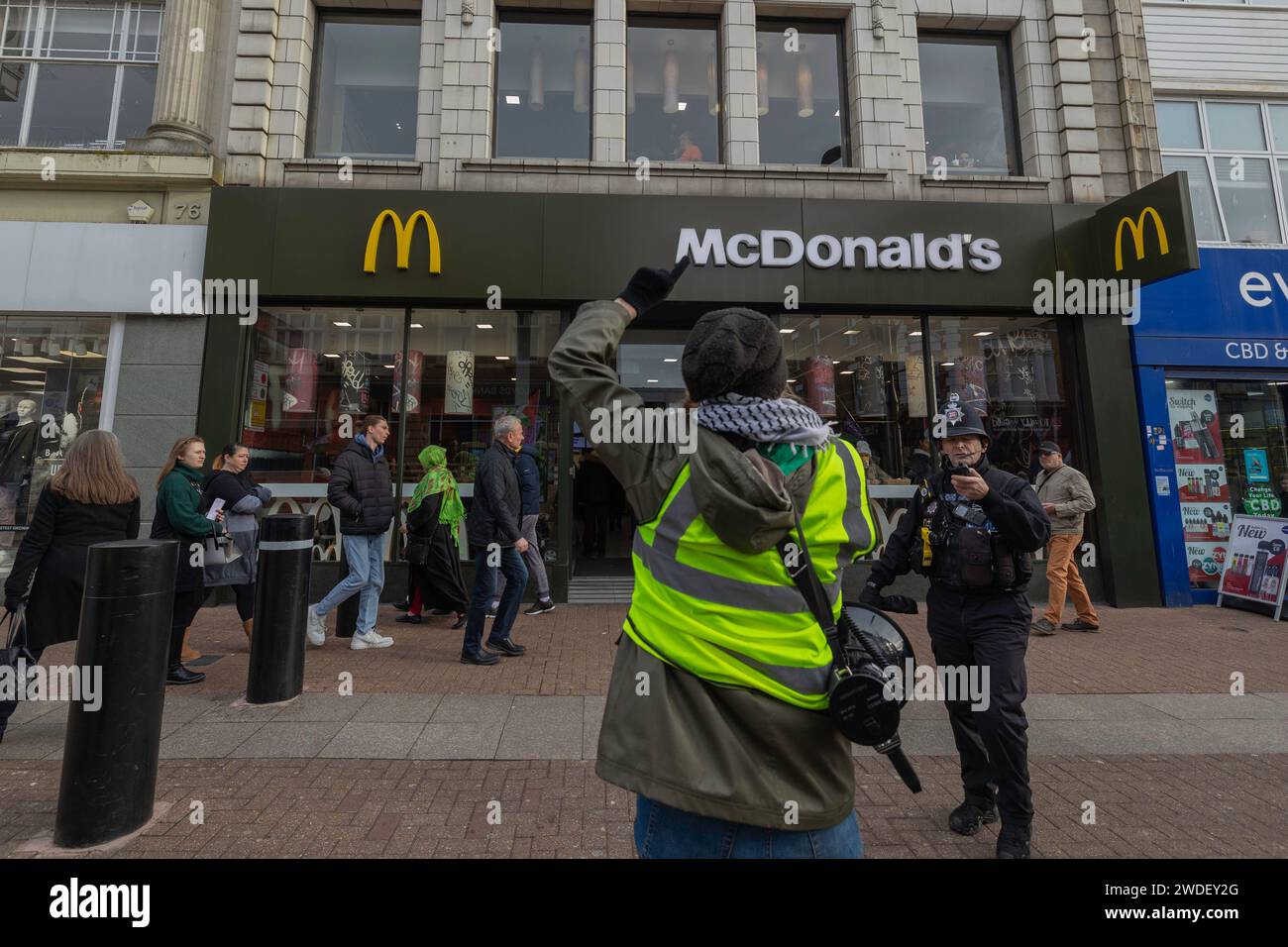 Southend on Sea, Regno Unito. 20 gennaio 2024. Un dimostratore che indossa un giubbotto ad alta visibilità solleva un braccio di fronte a McDonald’s durante una protesta per il cessate il fuoco in Palestina a Southend-on-Sea. Un agente di polizia si impegna con il manifestante mentre passano i pedoni. La scena cattura l'attivismo civico in un ambiente urbano commerciale, con segnaletica adiacente che fa riferimento ai prodotti CBD. La protesta chiede la fine della violenza a Gaza e la solidarietà con i civili palestinesi. I manifestanti organizzano una marcia e una piccola manifestazione per chiedere un cessate il fuoco in Palestina ora. Penelope Barritt/Alamy Live News Foto Stock