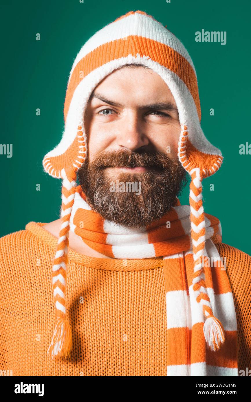 Ritratto in primo piano di un uomo attraente con barba e baffi in maglione arancione, divertente cappello e sciarpa a righe. Bell'uomo barbuto in abiti caldi Foto Stock