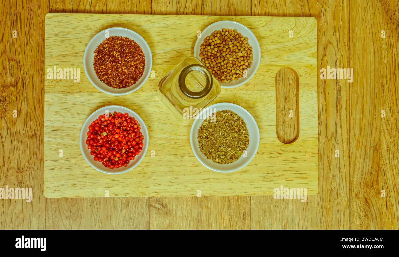 In alto a sinistra, pepe rosso macinato, coriandolo intero, pepe rosa e basilico in piccoli recipienti bianchi su un tagliere di legno con bottiglia di olio vuota Foto Stock