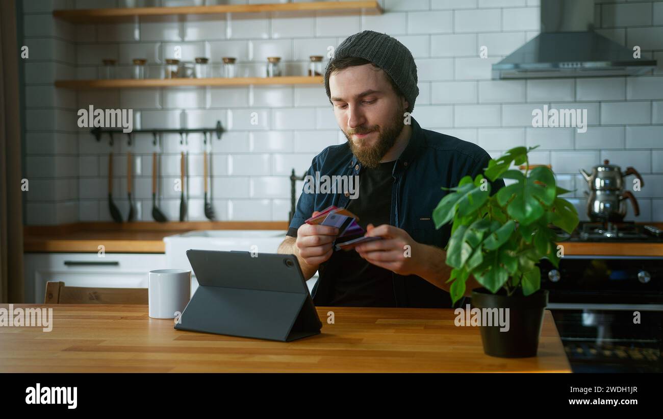 Un maschio con un berretto seduto in cucina a casa gli porta via le carte di credito e le prova una ad una per trovare la carta di credito che non ha Foto Stock