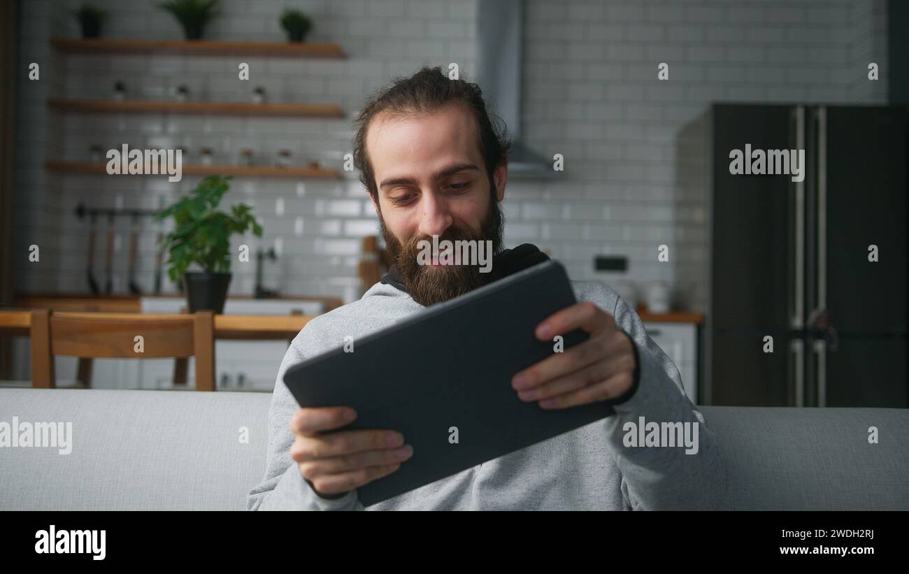 Giovane uomo barbuto con panino seduto sul divano a casa con sfondo in cucina, giocando emozionato concentrato coinvolto in un interessante gioco mobil Foto Stock