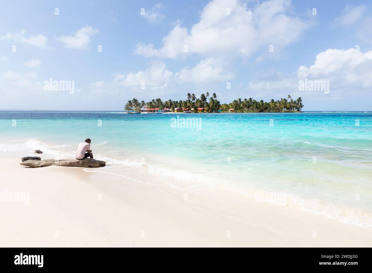Uomo del posto seduto su un tronco su una splendida spiaggia di sabbia con acqua turchese e un'isola tropicale sul retro, San blas, Mar dei Caraibi, Panama Foto Stock