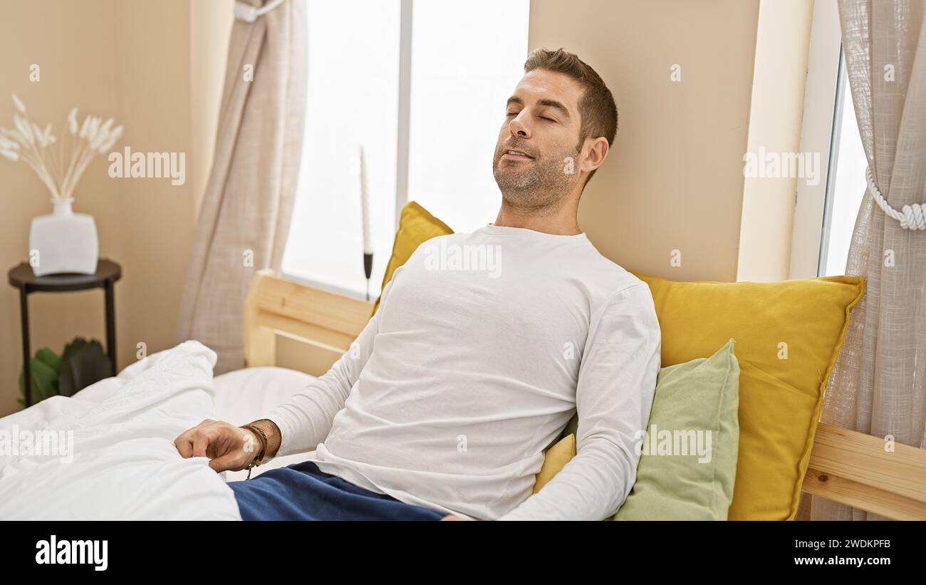 Un uomo rilassato con una barba lounge in una camera da letto accogliente, che evoca un senso di comfort e una casa moderna. Foto Stock