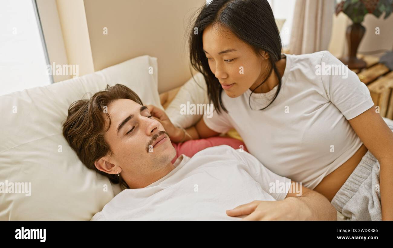 Coppia interrazziale che condivide un momento tenero in una camera da letto accogliente, con una donna che guarda amorevolmente un uomo sdraiato Foto Stock