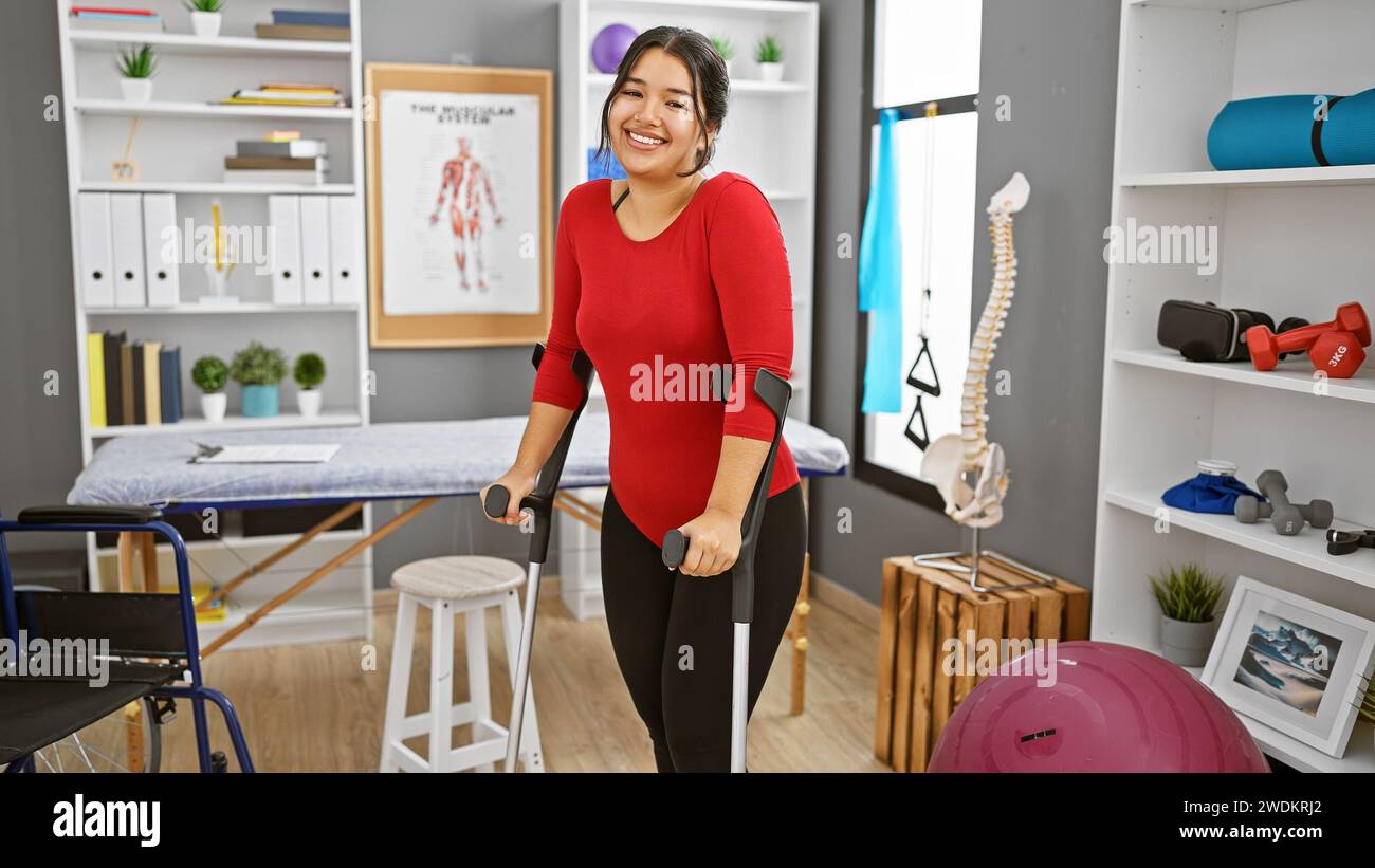 Una giovane donna ispanica sorridente che usa stampelle in una sala di terapia fisica ben attrezzata. Foto Stock