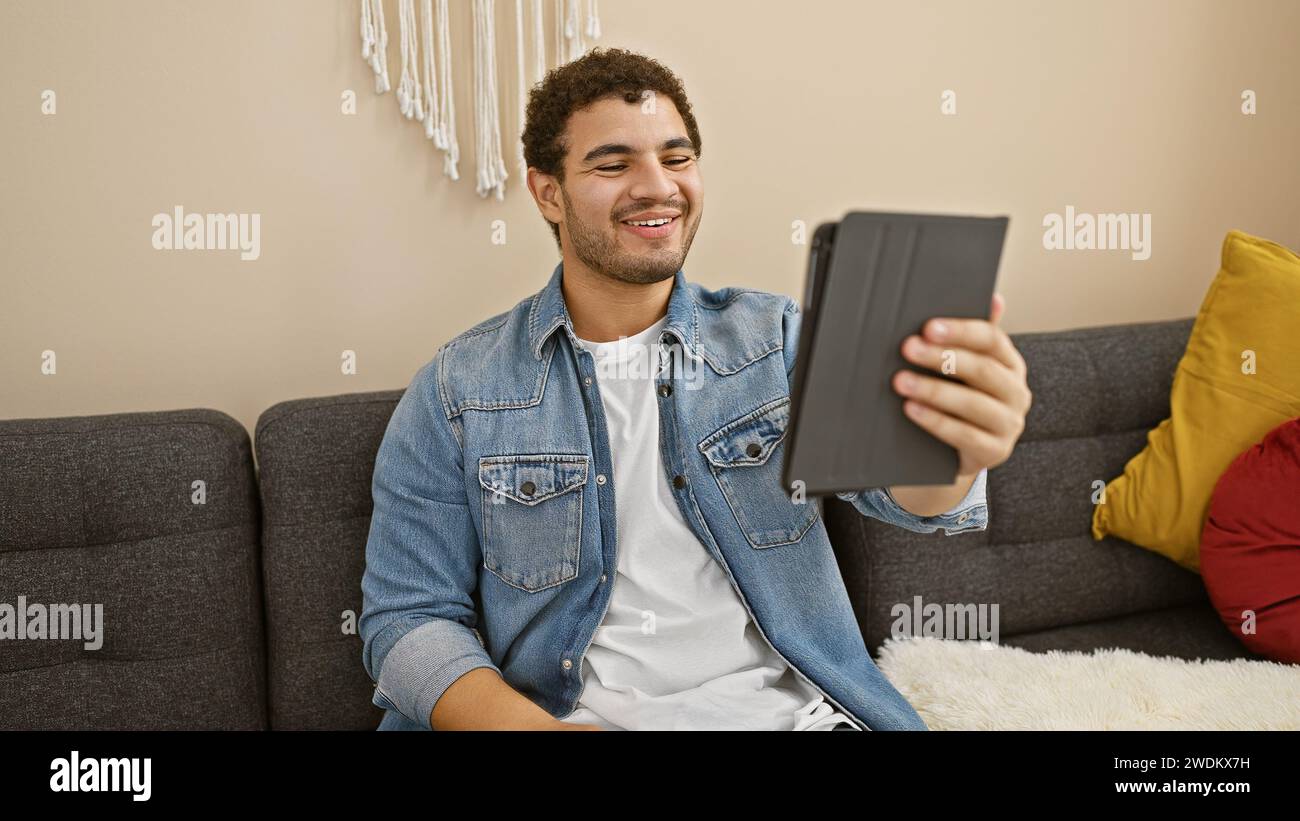 Un giovane sorridente con un tablet che si diverte a trascorrere il tempo libero su un divano in un accogliente salotto. Foto Stock