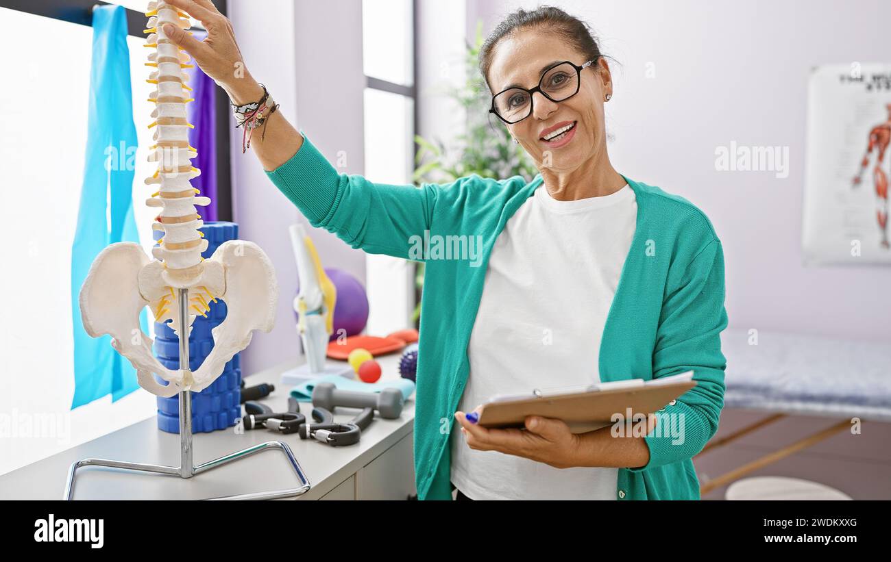 Una donna sorridente di mezza età con i capelli ricci spiega un modello spinale in una clinica di riabilitazione. Foto Stock