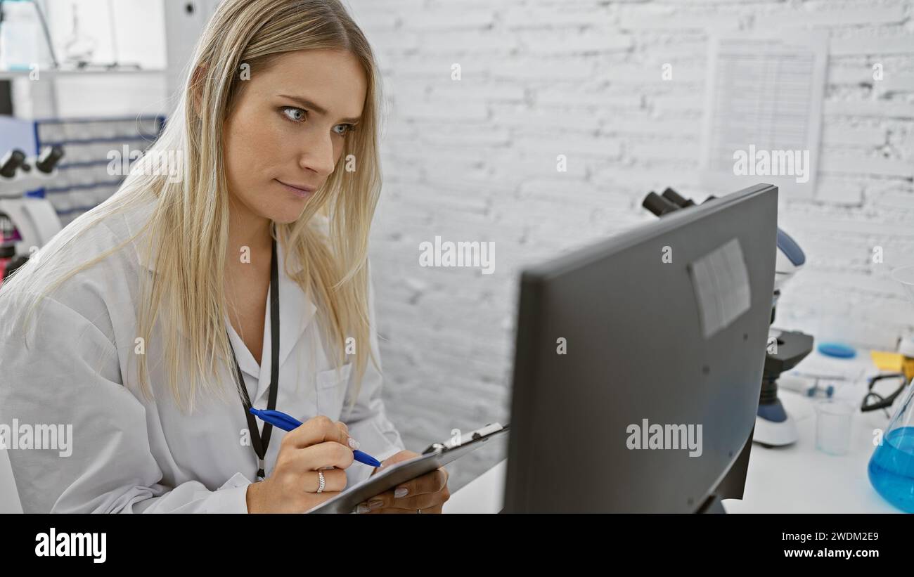 Attraente giovane scienziata bionda seriamente immersa nel lavoro di ricerca, utilizzando sapientemente la tecnologia informatica per l'analisi e la presa di appunti in una B. Foto Stock