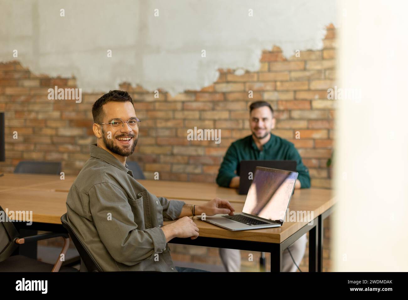 Due professionisti sorridenti si impegnano in una sessione di lavoro collaborativa su un tavolo di legno, il loro cameratismo è evidente in un ambiente da ufficio contemporaneo con un Foto Stock