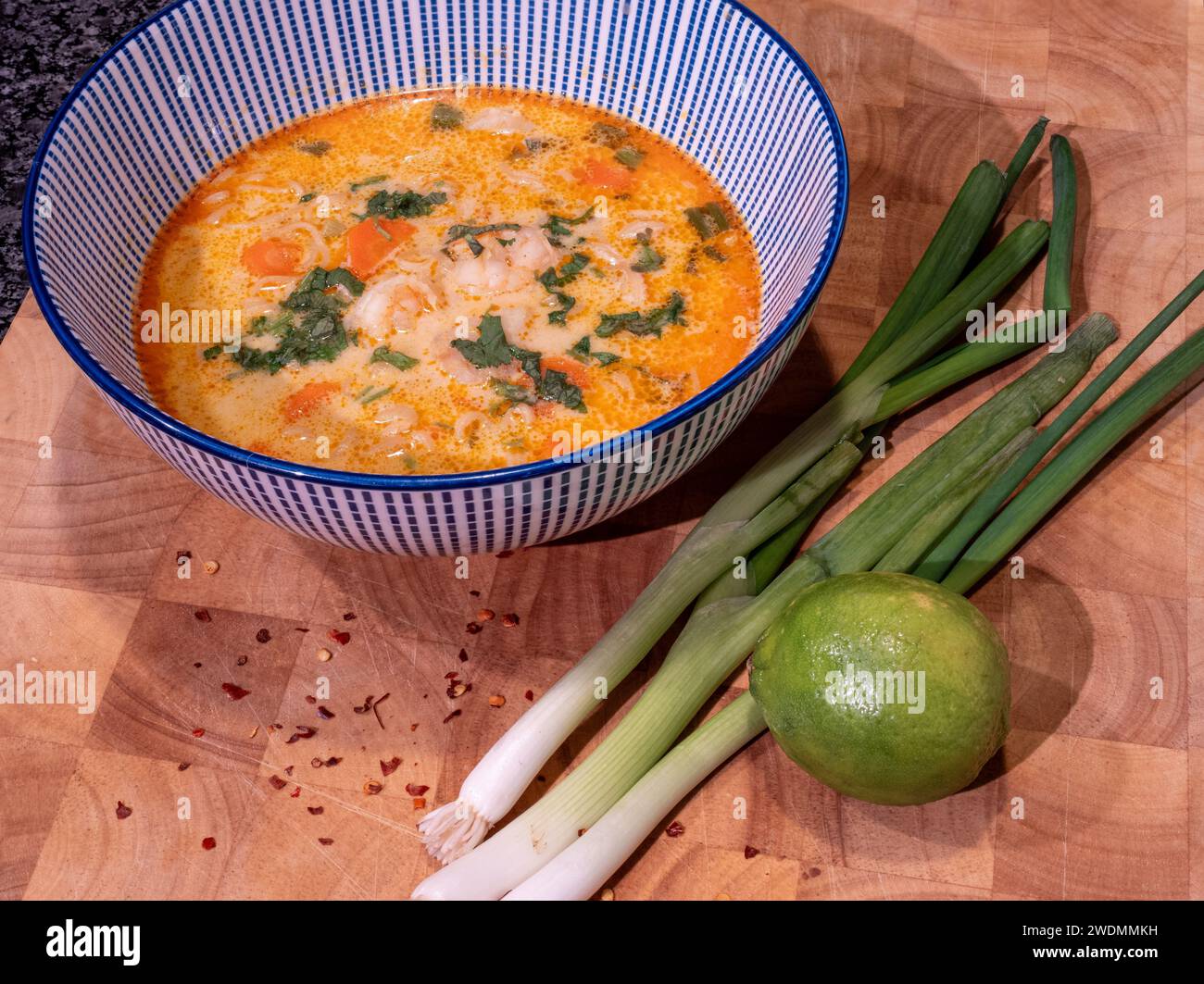 Assaporate i ricchi sapori di una zuppa di cocco asiatica infuso con pasta di curry rossa, elegantemente presentata in una ciotola blu sopra un blocco di legno. Accompagnato Foto Stock