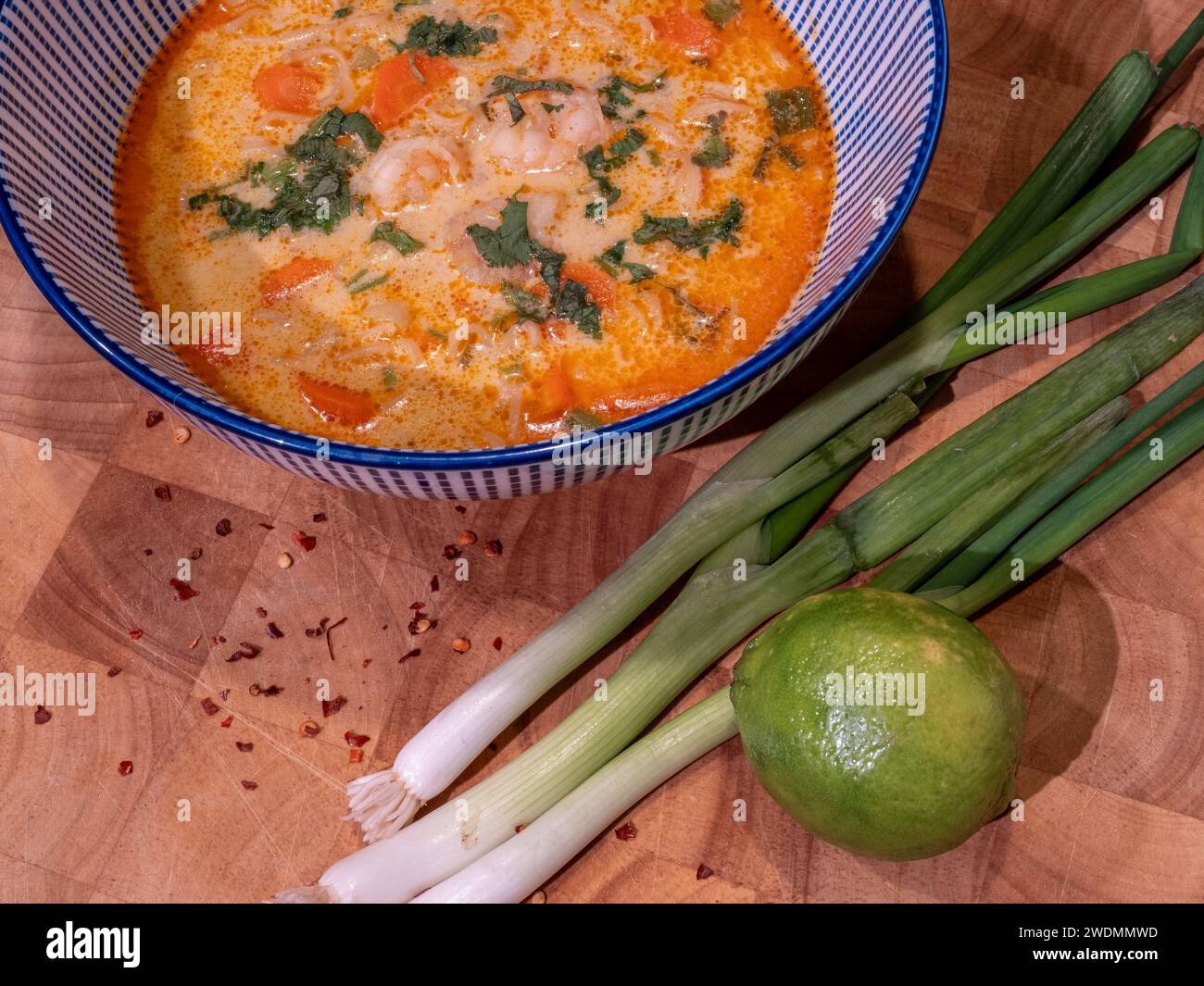 Assaporate i ricchi sapori di una zuppa di cocco asiatica infuso con pasta di curry rossa, elegantemente presentata in una ciotola blu sopra un blocco di legno. Accompagnato Foto Stock