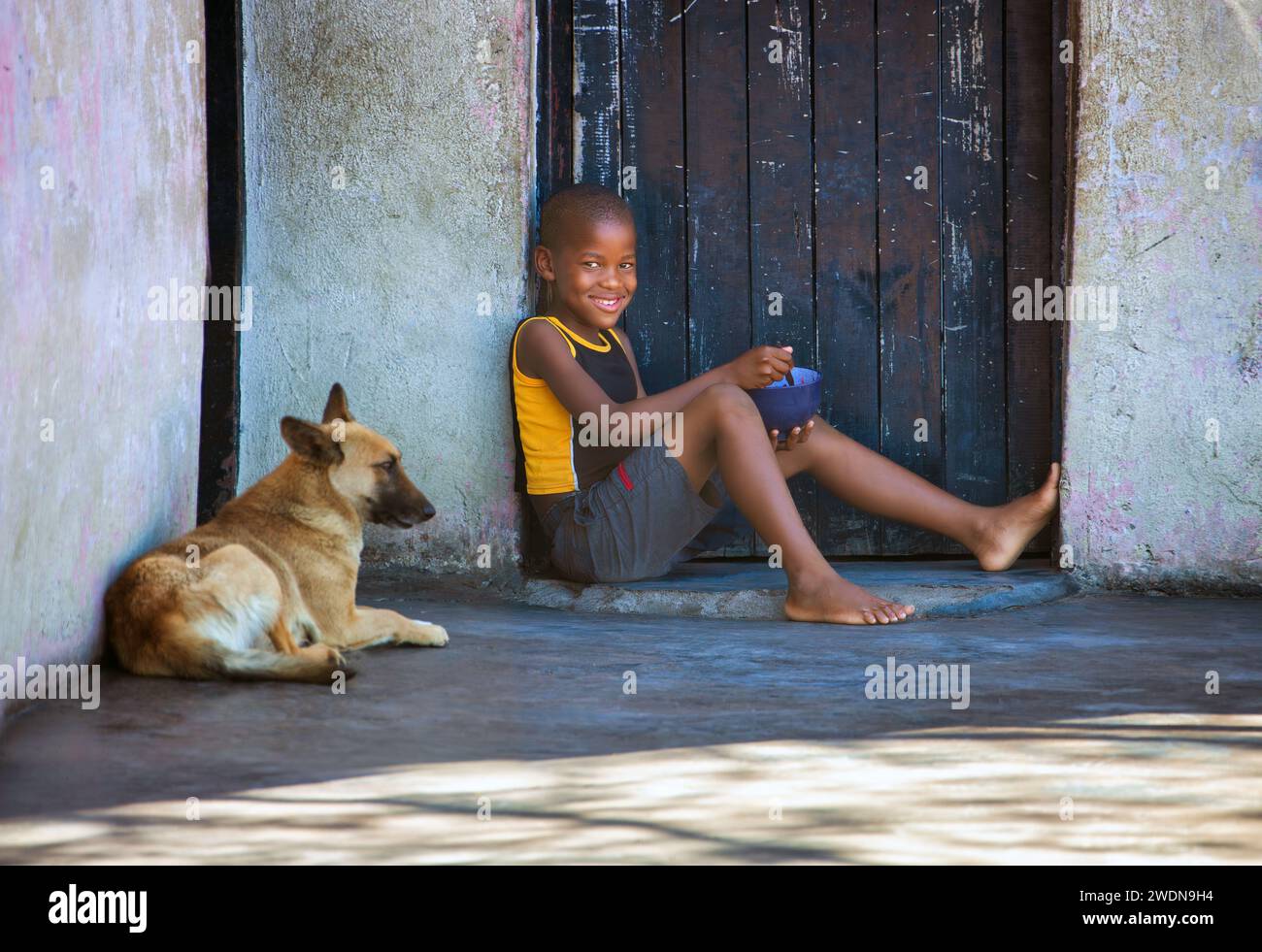 bambino africano affamato con il suo cane che pranza davanti alla porta Foto Stock