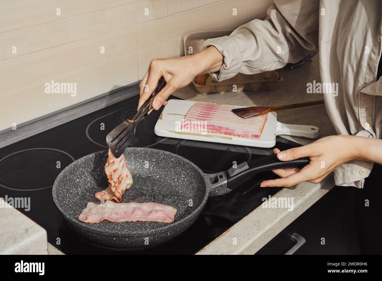 Mano di donna che capovolge il bacon sulla padella con le pinze da cucina Foto Stock