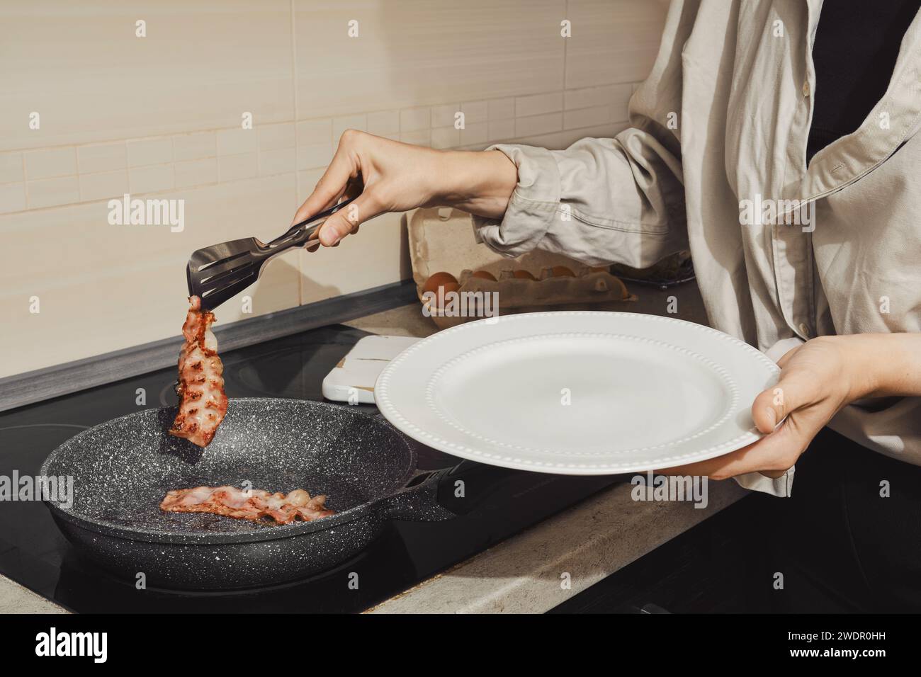Donna irriconoscibile che rimuove bacon fritto da una padella con pinze da cucina Foto Stock