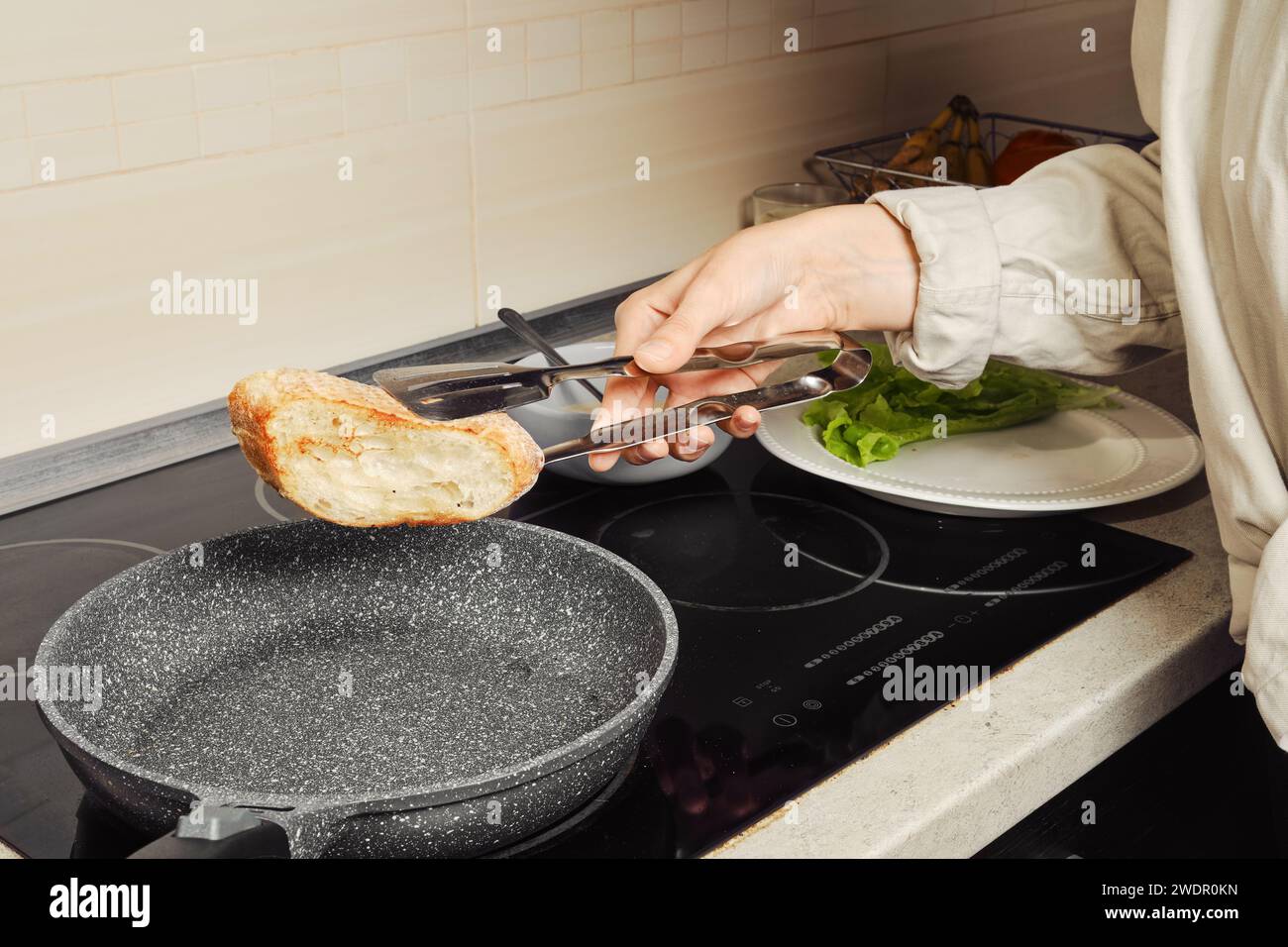 Donna irriconoscibile che gira un pezzo di pane tostato in una padella con pinze da cucina Foto Stock